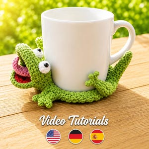 Könnte beinhalten: Eine weiße Keramiktasse steht auf einem grünen gehäkelten Alligator-Untersetzer. Der Alligator hat weiße Augen, eine rosa Zunge und einen roten Mund. Die Worte "Video Tutorials" werden darunter zusammen mit den Flaggen der USA, Deutschlands und Spaniens angezeigt.