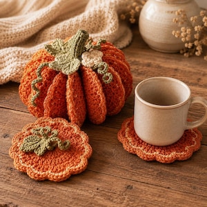 May include: A handcrafted orange crochet pumpkin with a green stem, leaves, and curly vines, accompanied by two matching round orange crochet coasters with green leaf accents, one of which supports a plain beige ceramic mug.