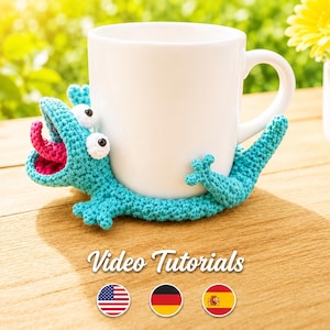 May include: A white ceramic mug rests on a turquoise crocheted lizard-shaped coaster with a pink tongue and white eyes. The image includes the text "Video Tutorials" and flags of the USA, Germany, and Spain.