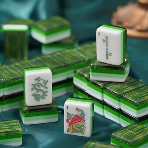 May include: A collection of rectangular mahjong tiles featuring a layered design with a white top section and a translucent green base. The tiles display engraved symbols including a green bamboo stalk, a green dragon, and a colorful bird illustration.
