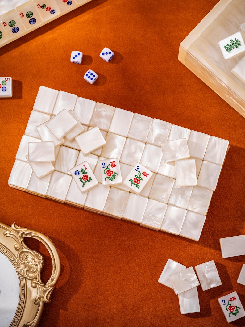 Puede incluir: Un juego de fichas de mahjong de color blanco nacarado con acabado liso. Tres fichas presentan dise&ntilde;os florales en rojo y verde con los n&uacute;meros 1, 2 y 3, y el texto AUT. Varias fichas en blanco est&aacute;n dispuestas en una cuadr&iacute;cula con tres dados.
