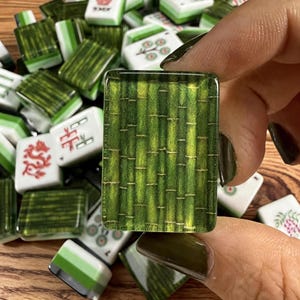 May include: A rectangular mahjong tile featuring a detailed green bamboo stalk pattern with a glossy finish. The tile is held against a pile of assorted mahjong tiles, some displaying white surfaces with red and green symbols and floral designs.