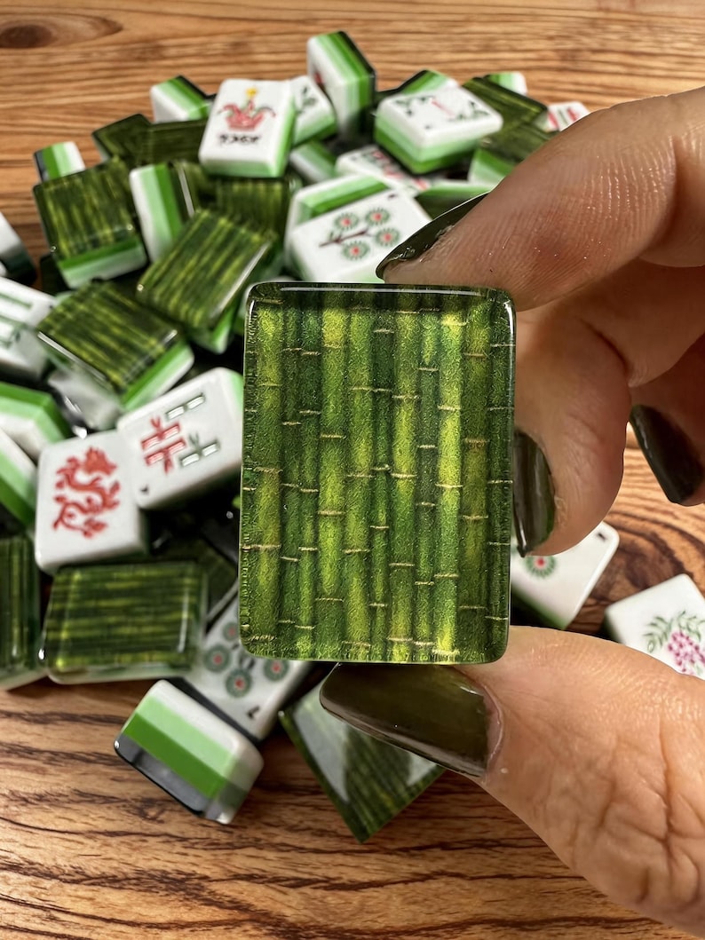 May include: A rectangular mahjong tile featuring a detailed green bamboo stalk pattern with a glossy finish. The tile is held against a pile of assorted mahjong tiles, some displaying white surfaces with red and green symbols and floral designs.