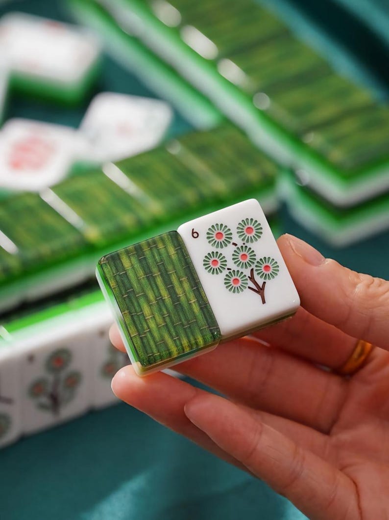 May include: A rectangular mahjong tile featuring a green bamboo stalk pattern on one side and a white surface with a floral design and the number 6 on the other. The tile is made of a smooth, glossy material with a distinct two-tone color scheme.