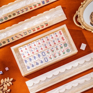 Puede incluir: Un juego de Mahjong que incluye una bandeja rectangular de madera llena de fichas color marfil marcadas con caracteres y s&iacute;mbolos chinos en rojo, azul y verde, junto a cuatro soportes largos para fichas con bordes festoneados.