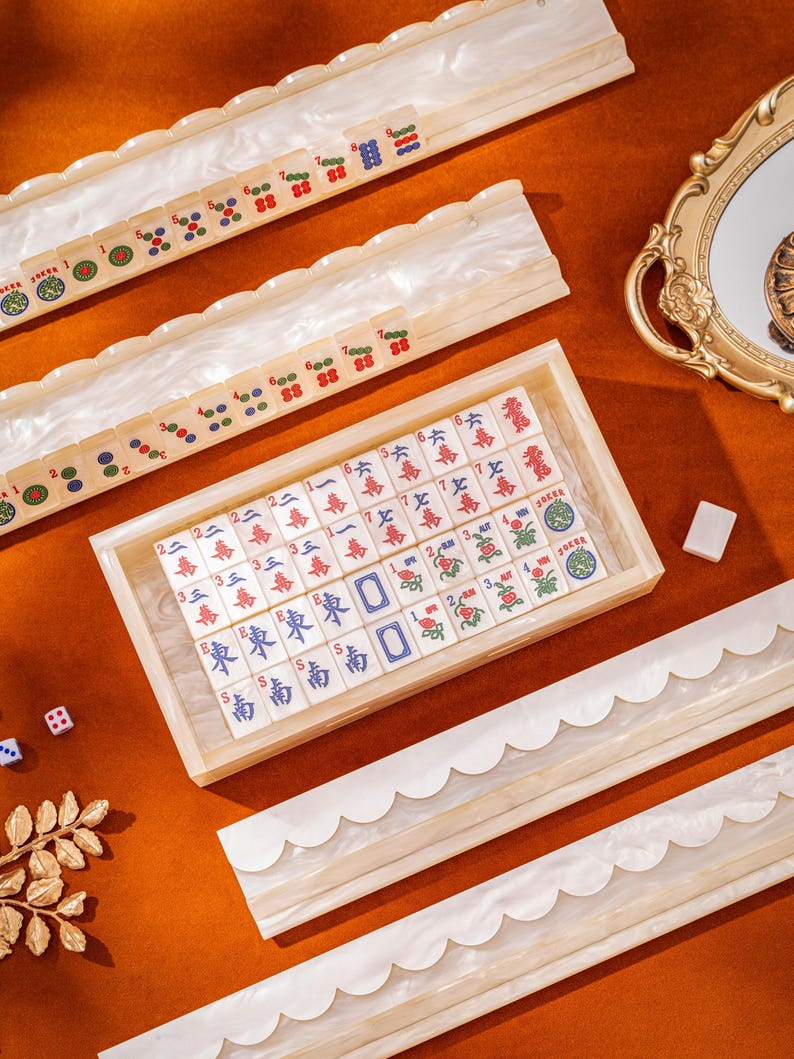 Puede incluir: Un juego de Mahjong que incluye una bandeja rectangular de madera llena de fichas color marfil marcadas con caracteres y s&iacute;mbolos chinos en rojo, azul y verde, junto a cuatro soportes largos para fichas con bordes festoneados.