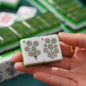 May include: Two rectangular Mahjong tiles with white faces and green backs. Each tile features a printed design of a stylized tree with pink and green circular blossoms. The number 6 is printed in black on the top left corner of each tile.