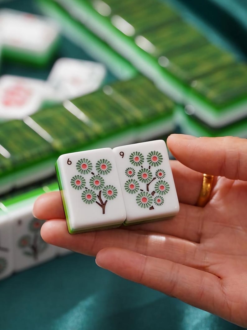 May include: Two rectangular Mahjong tiles with white faces and green backs. Each tile features a printed design of a stylized tree with pink and green circular blossoms. The number 6 is printed in black on the top left corner of each tile.