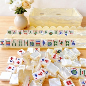 Puede incluir: Un juego de mahjong con fichas de color blanco nacarado que presentan s&iacute;mbolos, n&uacute;meros y caracteres grabados en color. El set incluye dos soportes festoneados, una caja de almacenamiento a juego y fichas sueltas sobre una superficie de madera.