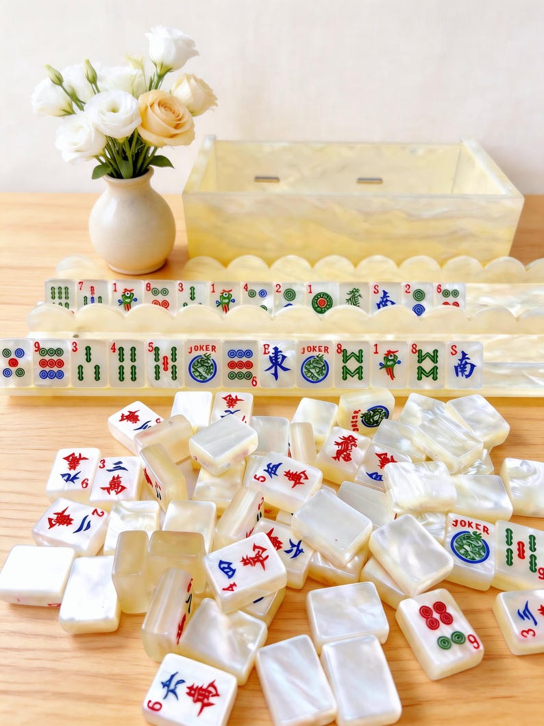 Puede incluir: Un juego de mahjong con fichas de color blanco nacarado que presentan s&iacute;mbolos, n&uacute;meros y caracteres grabados en color. El set incluye dos soportes festoneados, una caja de almacenamiento a juego y fichas sueltas sobre una superficie de madera.