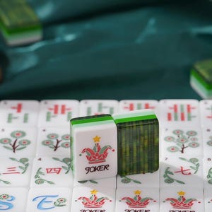 May include: A set of white mahjong tiles featuring colorful floral and character designs. Two tiles stand upright in the center, one displaying a red and green jester hat icon with a yellow star and the word JOKER, and the other showing a bamboo pattern.