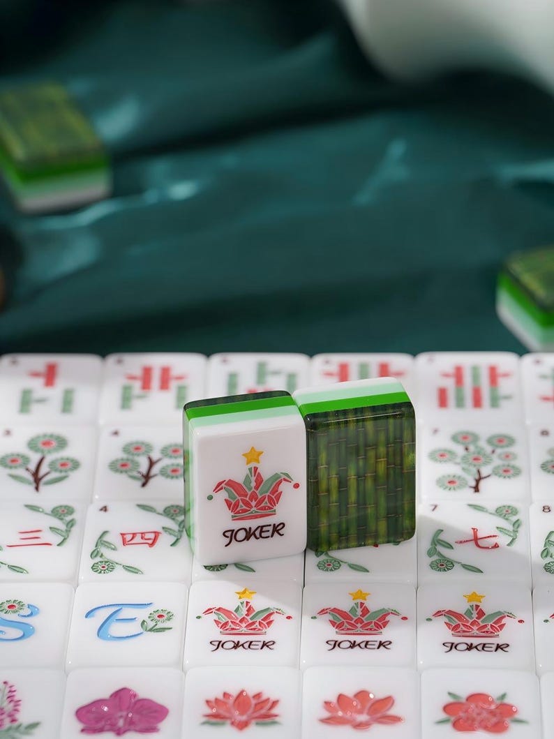 May include: A set of white mahjong tiles featuring colorful floral and character designs. Two tiles stand upright in the center, one displaying a red and green jester hat icon with a yellow star and the word JOKER, and the other showing a bamboo pattern.