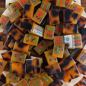 May include: A collection of rectangular mahjong tiles with a translucent amber and dark brown tortoiseshell pattern. The tiles feature engraved symbols, numbers, and characters in red, green, and black ink, including a Joker tile with a dragon design.