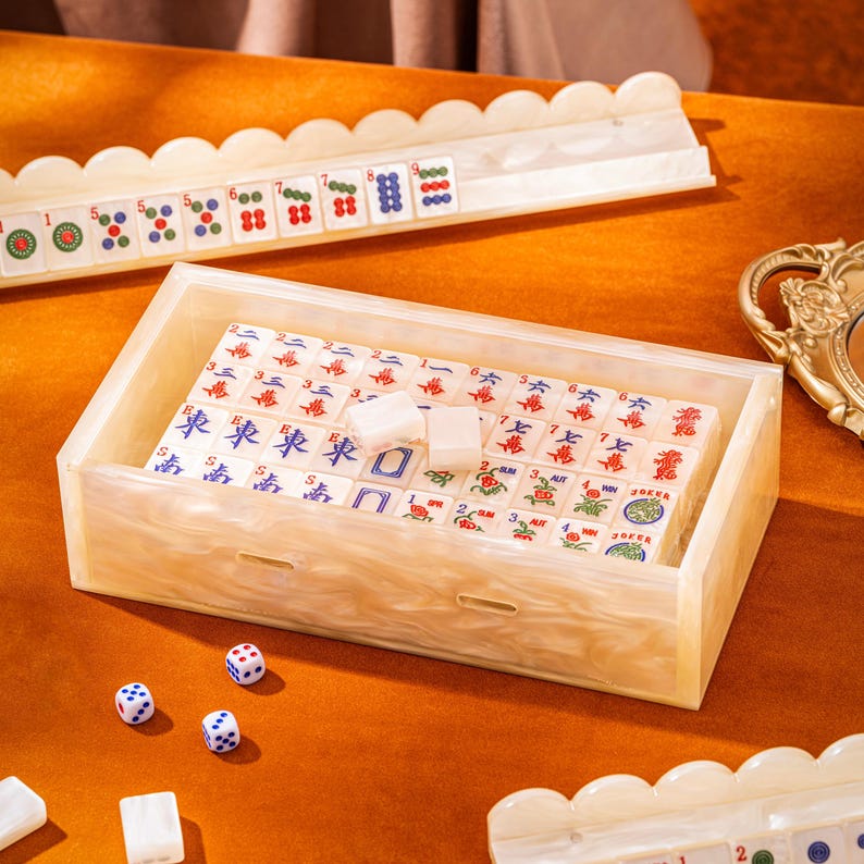 Puede incluir: Una caja rectangular para mahjong con acabado en crema marmoleado que contiene numerosas fichas blancas con marcas de caracteres y palos en rojo, azul y verde. El juego incluye soportes para fichas a juego con bordes festoneados y tres dados blancos.