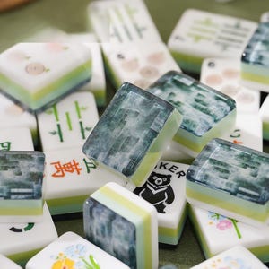 May include: A collection of rectangular mahjong tiles featuring a layered design with a white top, a light green middle, and a dark green base. The tiles display various illustrations including floral patterns, bamboo stalks, and a black bear graphic.