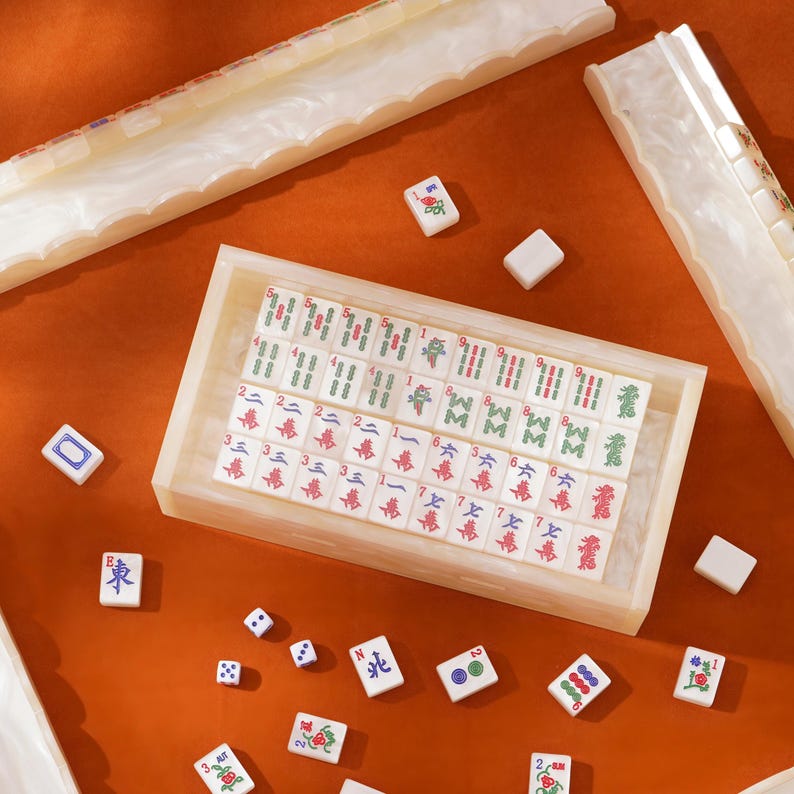 Puede incluir: Un juego de mahjong con fichas blancas que presentan caracteres y s&iacute;mbolos en rojo, verde y azul. El conjunto incluye una caja de almacenamiento rectangular, soportes para fichas y dados. Las fichas est&aacute;n dispuestas en filas dentro de la caja, con otras fichas sueltas y dados esparcidos sobre una superficie naranja.