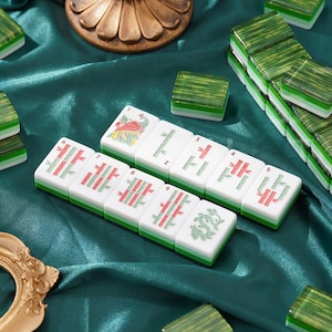 May include: A set of rectangular mahjong tiles featuring green bamboo patterns, a red bird, and a green dragon design. The tiles have white faces with green and white layered bases, arranged in rows alongside other scattered tiles on a dark green fabric.