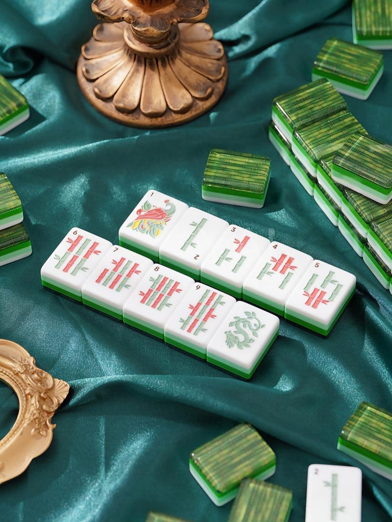 May include: A set of rectangular mahjong tiles featuring green bamboo patterns, a red bird, and a green dragon design. The tiles have white faces with green and white layered bases, arranged in rows alongside other scattered tiles on a dark green fabric.