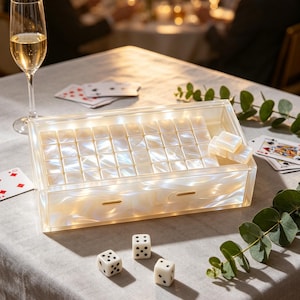 May include: A clear, rectangular game box with a pearlized interior, illuminated by internal lights. Three white dice with black dots are scattered on the table. A glass of champagne and playing cards are also visible, suggesting a game night setting.