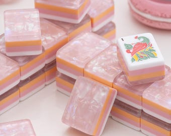 Peut inclure: Une collection de tuiles de mah-jong rectangulaires présentant un design en couches avec un dessus rose nacré translucide, une bande centrale orange, une bande rose et une base blanche. Certaines tuiles arborent des motifs d'oiseaux ou de dragons.