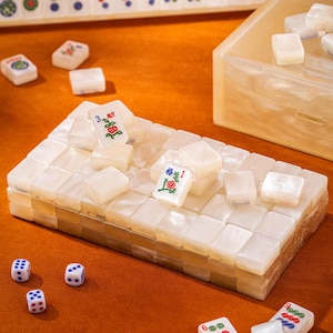 Puede incluir: Un conjunto de fichas de mahjong de color blanco nacarado dispuestas en una pila rectangular, con fichas individuales que presentan dise&ntilde;os florales rojos y verdes y los n&uacute;meros 1 y 3. Tres dados blancos con puntos azules y rojos est&aacute;n colocados cerca.