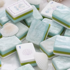 May include: A collection of rectangular mahjong tiles featuring a marbled light blue and white surface with green and white layered bases. Some tiles display printed illustrations of seashells and the number 1, interspersed with small white seashells.