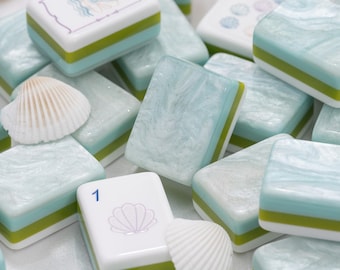 Puede incluir: Una colección de fichas de mahjong rectangulares con un acabado jaspeado en azul claro y blanco sobre bases de capas verdes y blancas. Algunas fichas muestran iconos de conchas impresos y el número 1, entremezcladas con pequeñas conchas.