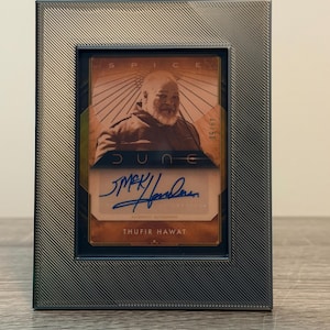 May include: A collectible Dune trading card featuring an image of Thufir Hawat with a blue ink autograph. The card is encased in a dark gray, textured frame with diagonal lines. The text on the card includes SPICE, DUNE, and THUFIR HAWAT.