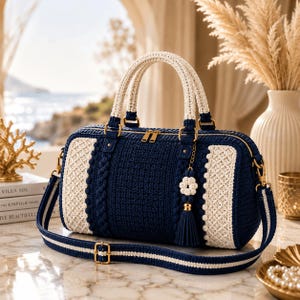 May include: A navy blue and cream crochet handbag featuring a structured rectangular body, two top handles, and a detachable shoulder strap. The bag includes a gold-tone zipper closure and a decorative flower-shaped tassel charm hanging from the side.
