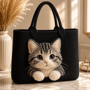 Peut inclure: Un sac fourre-tout noir en crochet orné d'une application texturée en trois dimensions représentant un chat tigré gris et blanc. Le motif du chat comprend des moustaches détaillées, de grands yeux sombres et deux pattes avant.