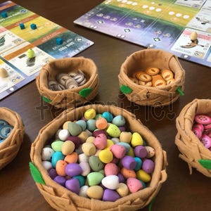 May include: Five tan felt baskets shaped like bird nests, each featuring small green felt leaves. Four baskets contain round game tokens with blue fish, orange seeds, gray rodents, and pink berries. The center basket holds many small, colorful eggs.