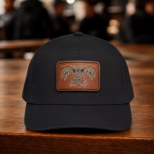 Puede incluir: Una gorra de béisbol negra con un parche rectangular de cuero marrón en la parte delantera. El parche muestra una ilustración grabada de dos criaturas con cuernos estilizadas frente a una motocicleta, con el texto gnu heart me debajo.