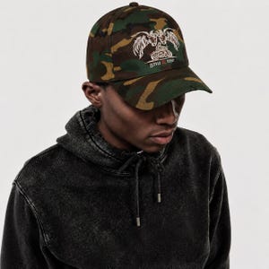 May include: A camouflage baseball cap featuring an embroidered graphic of two winged creatures flanking a motorcycle, with the text gnu & me printed below in a clean font. The cap has a structured crown, a curved brim, and a button on top.