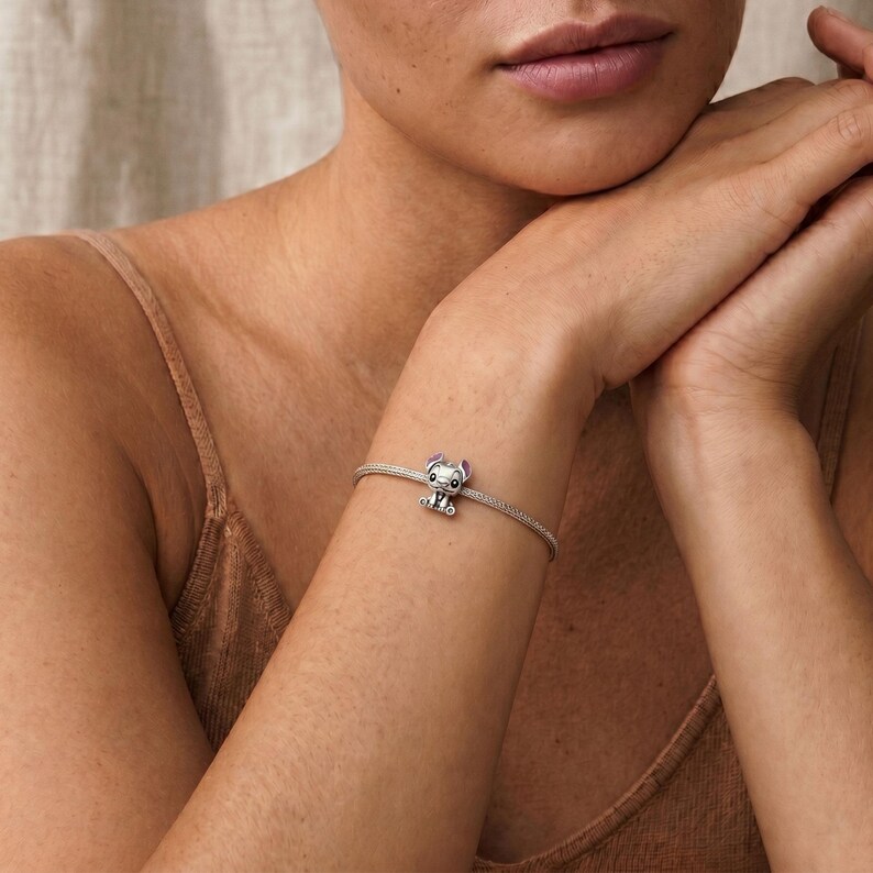 May include: A silver-toned charm bracelet featuring a small, detailed character charm with large black eyes and light purple inner ears. The charm is centered on a thin, flexible, woven metal chain that wraps around the wrist.