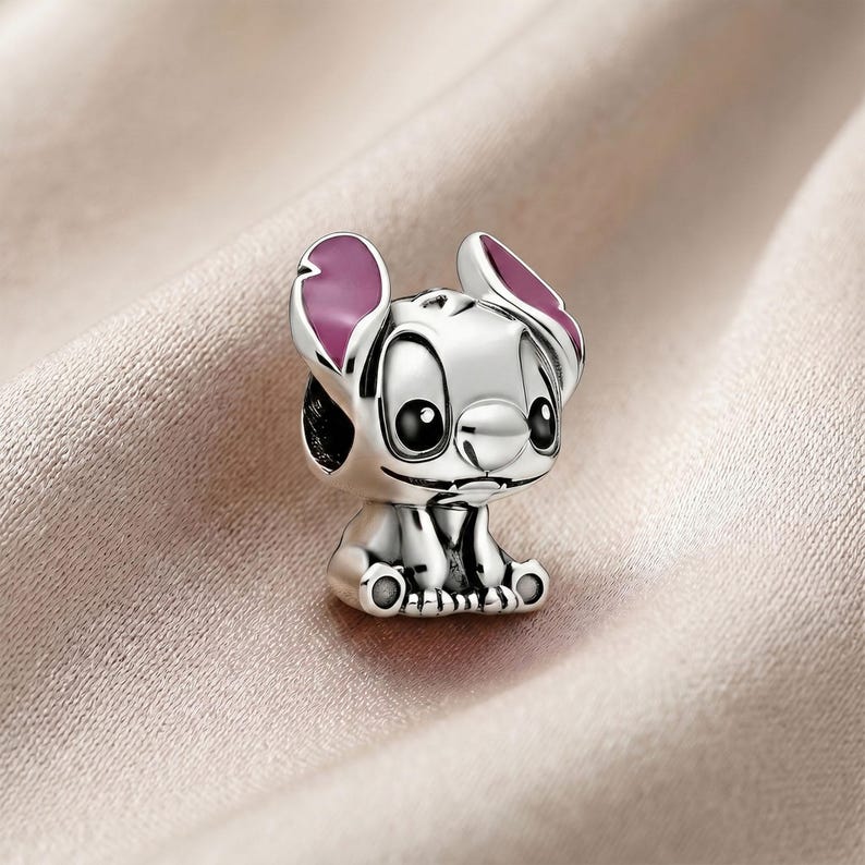 May include: A silver-toned metal charm shaped like a cartoon character with large eyes, a small nose, and prominent ears featuring pink enamel accents on the inner surfaces. The character is depicted in a seated position with visible paws and feet.