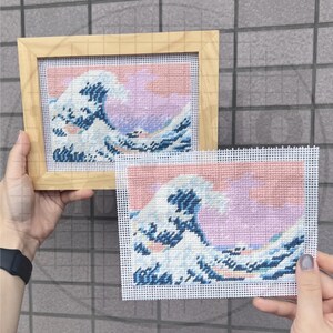 May include: Needlepoint art featuring a stylized wave design in shades of blue, white, and pink. One piece is framed in a light wood frame, while the other is unframed. The artwork is inspired by Japanese art.