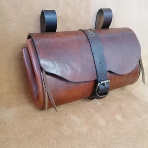Puede incluir: Un bolso cilíndrico de cuero marrón con un cierre de solapa liso asegurado por una correa de cuero marrón oscuro con una hebilla de metal. Dos trabillas de cuero marrón oscuro están unidas a la parte superior para su montaje y los bordes están cosidos.