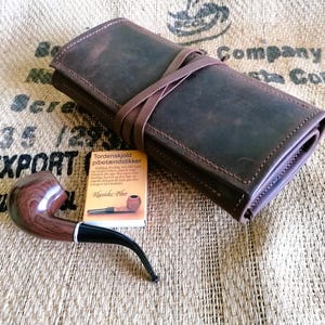 May include: A dark brown leather roll-up case with a wrap-around strap, a polished wooden tobacco pipe with a black stem, and a small rectangular box of Tordenskjold pibetændstikker matches with a printed image of a pipe on the front label.