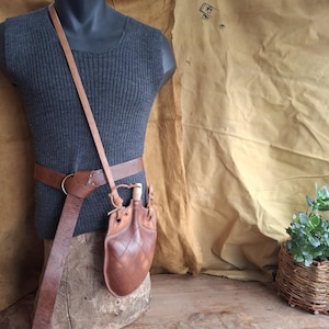 Peut inclure: Une gourde en cuir marron avec un motif surpiqué en losange et un bouchon en bois clair, suspendue à une longue et fine bandoulière en cuir et fixée à une large ceinture en cuir marron avec une boucle en métal en forme d'anneau.