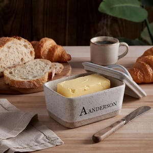 Puede incluir: Una mantequillera rectangular de cerámica con acabado crema moteado y el texto A ANDERSON impreso en el frente. El recipiente contiene un bloque de mantequilla amarilla y tiene una tapa a juego apoyada entreabierta al lado sobre una superficie de madera.
