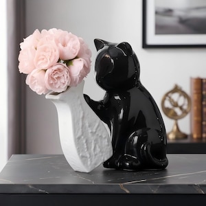 Könnte beinhalten: Eine glänzend schwarze Keramikkatzenfigur, die eine weiße, texturierte Keramikvase hält. Die Vase enthält einen Strauß künstlicher blassrosa Rosen. Die Katze ist in sitzender Position dargestellt, wobei eine Vorderpfote am Vasenrand ruht.