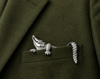 May include: A silver-toned metal alligator brooch attached to the breast pocket of an olive green blazer. The alligator figure features detailed scales, a textured snout, and visible claws, with its tail extending over the edge of the pocket.