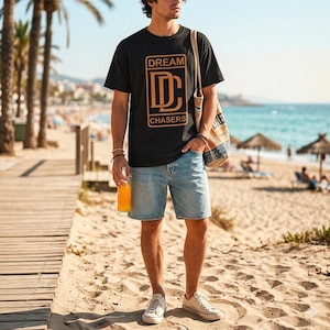 May include: Black t-shirt with the text "DREAM CHASERS" in a gold rectangular design. The person is wearing light blue denim shorts and white sneakers. A canvas tote bag is slung over the shoulder. The background shows a beach with palm trees and the ocean.