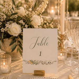 Peut inclure: Une carte de numéro de table rectangulaire blanche avec le mot Table et le chiffre 1 dans une police cursive sombre. La carte présente une illustration botanique horizontale de feuilles vertes, de petits citrons jaunes et d'olives en bas.