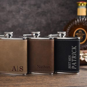 May include: Three stainless steel flasks with leather-effect wraps in tan, brown, and black. The tan flask features the initials A|S, the brown flask is engraved with Nathan and a date, and the black flask displays the text BEST MEN PATRICK.