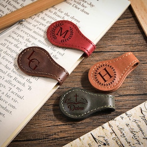 May include: Four teardrop-shaped leather magnetic bookmarks in red, dark brown, tan, and olive green. Each features a laser-engraved design: a laurel wreath with the letter M, the letters LG, a laurel wreath with the letter H, and a cross with the name Daine.