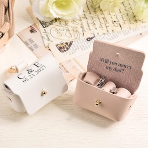 May include: Two small rectangular leather ring boxes with gold-tone snap closures. One white box is closed and engraved with C & E 05.21.2027. One light pink box is open, revealing internal ring slots and the text Will you marry my dad?