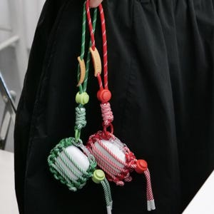 May include: Two white earbud cases encased in woven rope nets, one in green and one in red. Each features a matching colored cord lanyard with a small wooden tag labeled "innerz," a sliding plastic toggle, and white cord ends for secure carrying.