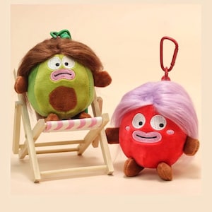 May include: Two plush character keychains with hair. One is a green avocado-shaped figure with brown hair sitting on a miniature wooden beach chair. The other is a red round figure with lavender hair and a red carabiner clip attached to the top.