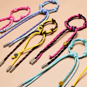 May include: A collection of lanyards made from colorful braided cord with reflective white stitching. Each lanyard features a circular knotted loop at the top, metallic silver end caps, and small circular smiley face charms in yellow or silver.